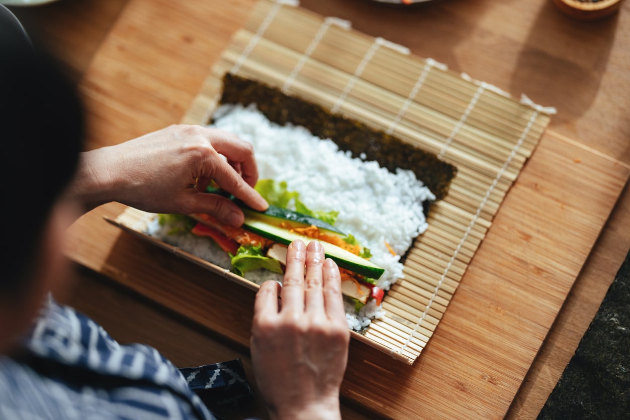 Hands placing cucumber slices onto sushi rice and fillings spread on a bamboo rolling mat.
