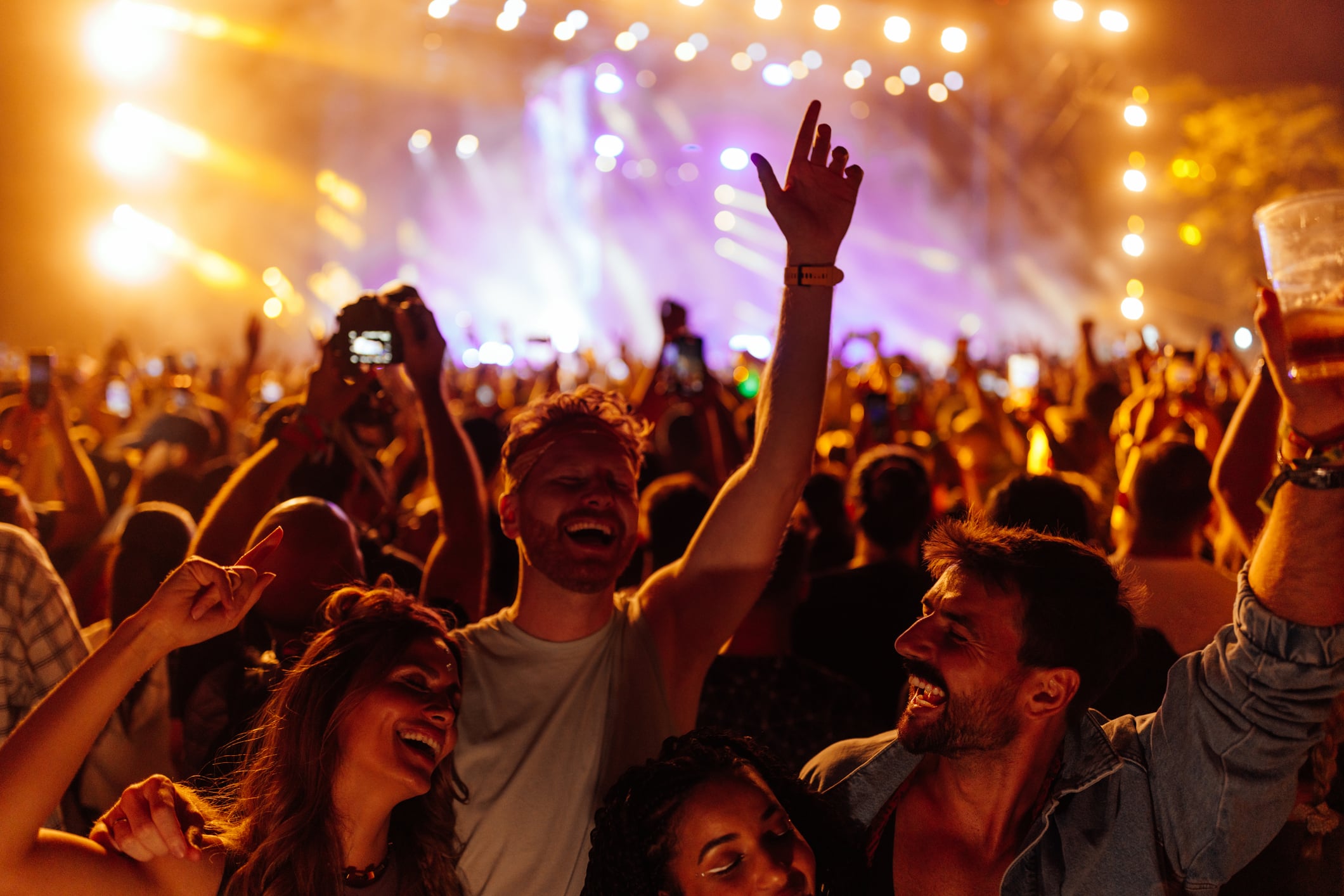 A diverse group of people dancing and cheering at a crowded outdoor concert with bright stage lights in the background.