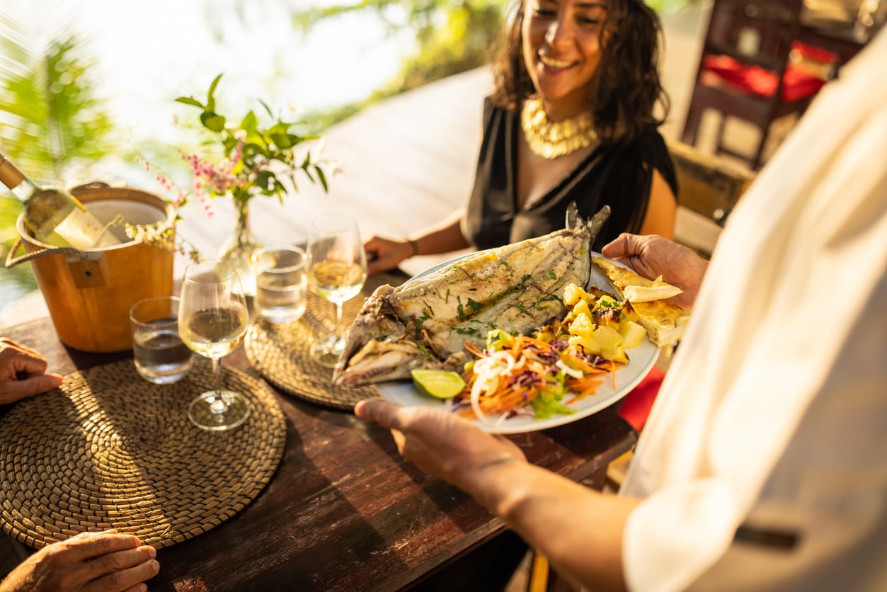 A server places a plate of grilled whole fish and vegetables—a popular restaurant menu tip for healthy dining—in front of a smiling woman at an outdoor table set with drinks.