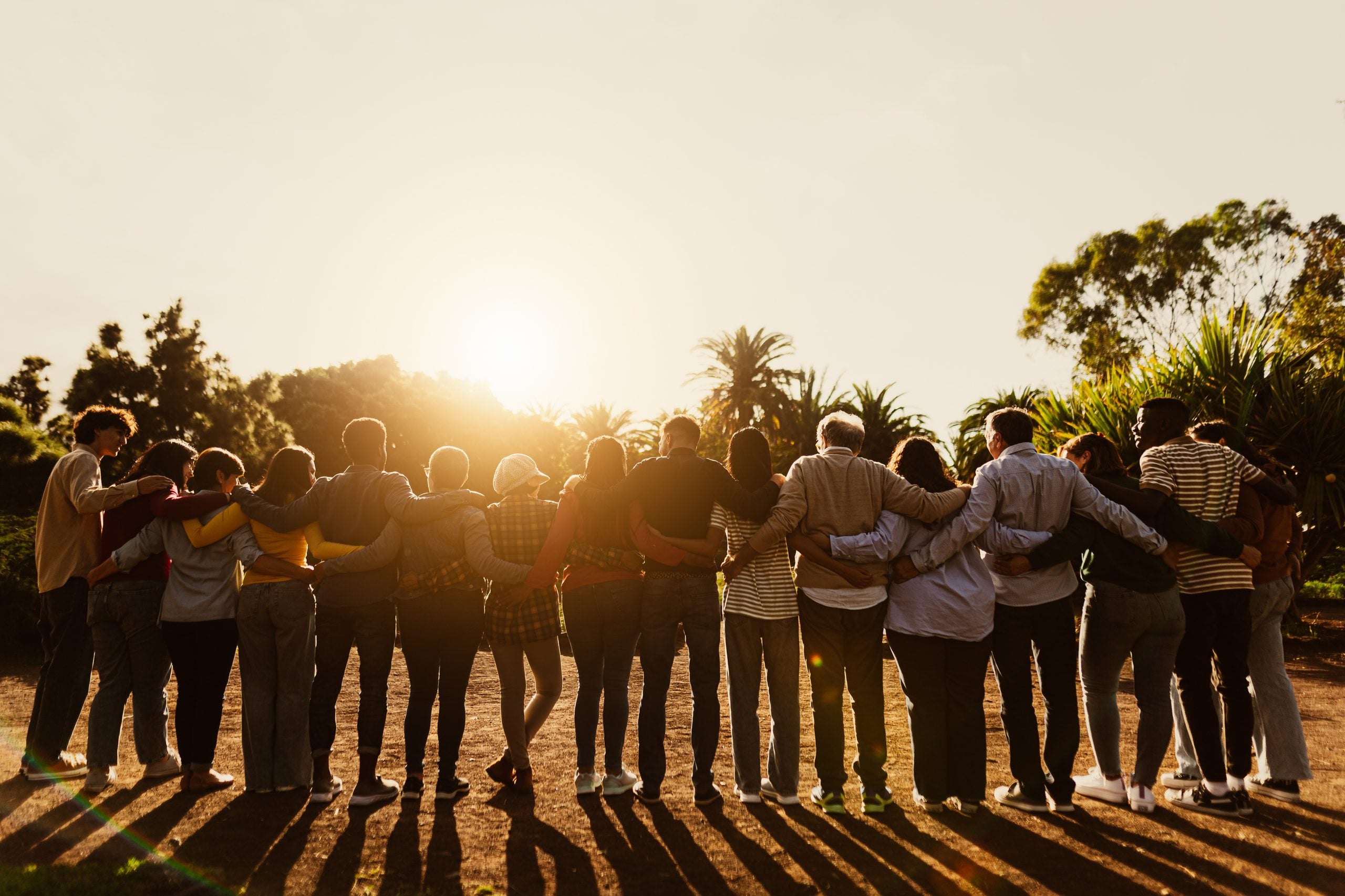 A large group of people standing in a row with their arms around each other, silhouetted against a setting sun in a park.