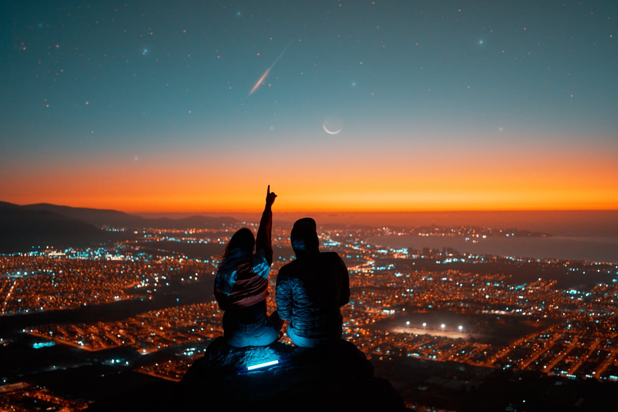 Two people sit on a hilltop overlooking a city at night, with one person pointing toward a shooting star in the sky above a crescent moon.