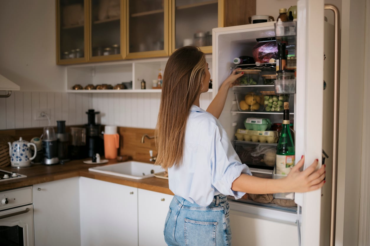 A woman in a blue shirt standing in a kitchen reaches into an open refrigerator to retrieve a food container.