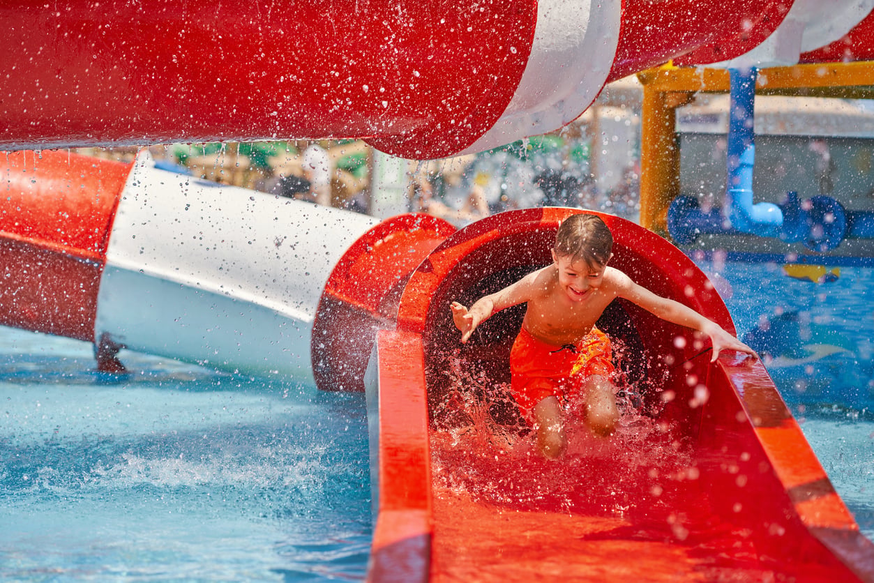 A boy in orange swim trunks slides down a red water slide, splashing into the pool during cruise entertainment.