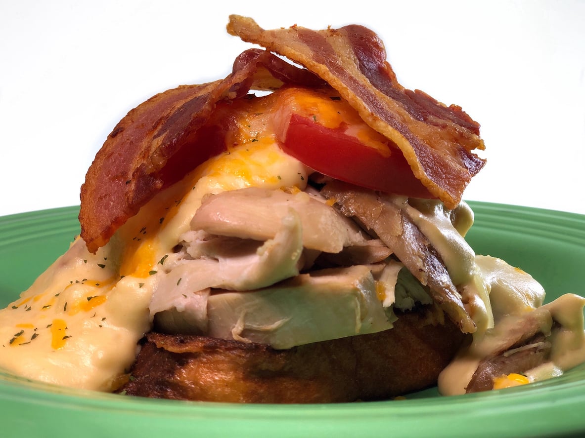 Open-faced hot turkey sandwich topped with melted cheese, tomato slices, and bacon on a green plate.