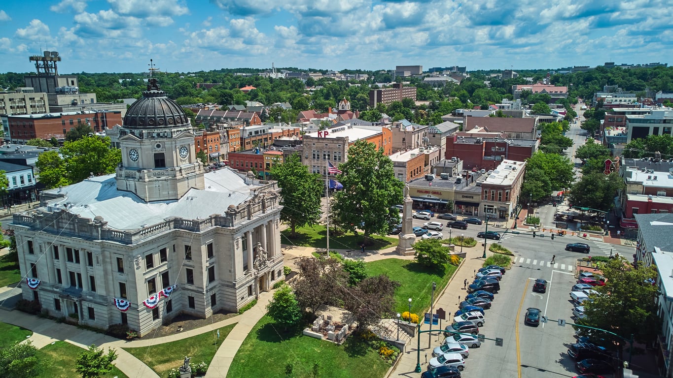 Aerial view of a town square featuring a historic stone courthouse with a dark dome and surrounding city buildings, streets, and parked cars.