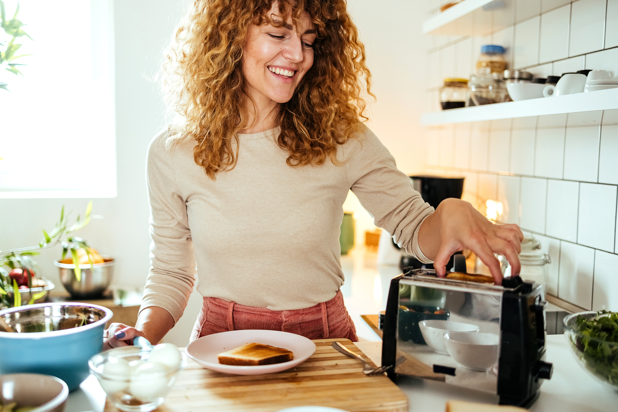 A woman with curly red hair smiles while pressing down the lever on a toaster in her kitchen.