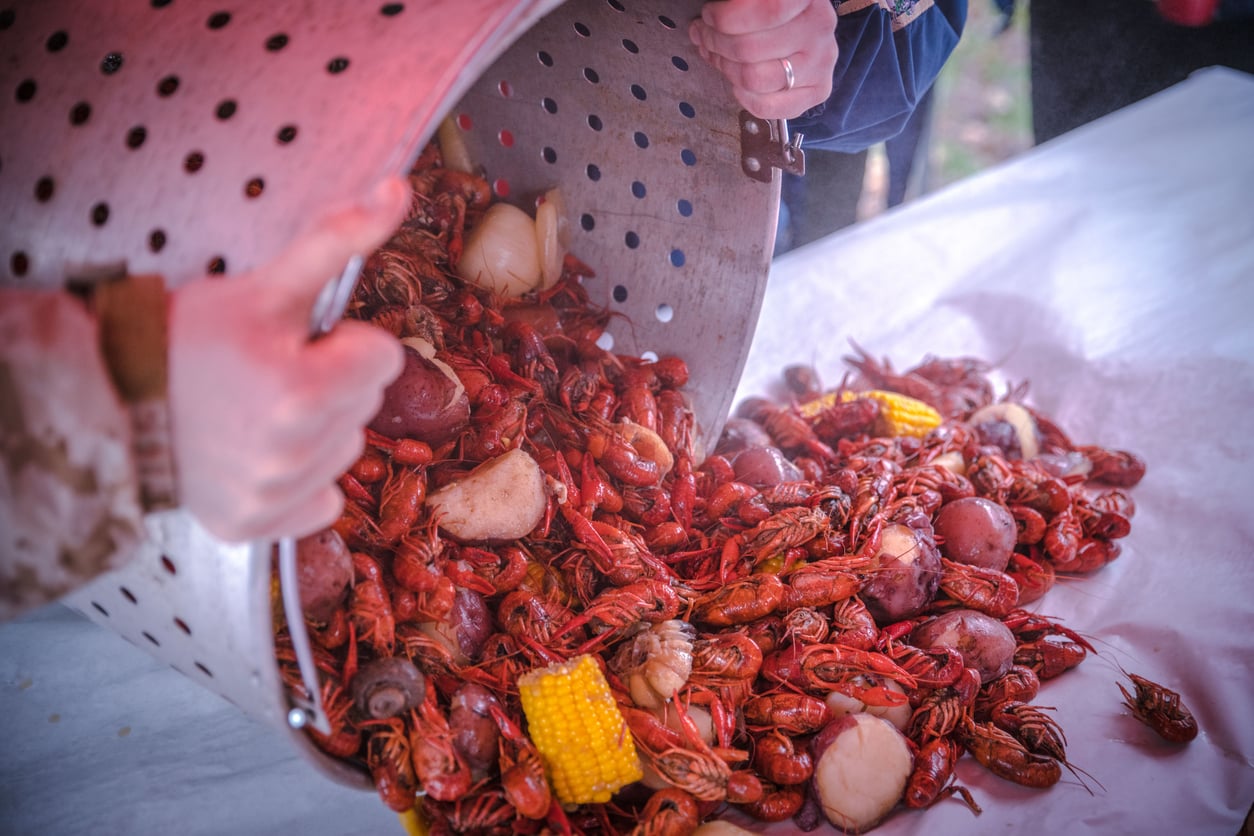 Hands dump a large, perforated metal pot filled with boiled crawfish, red potatoes, onions, and corn onto a table covered with white paper.