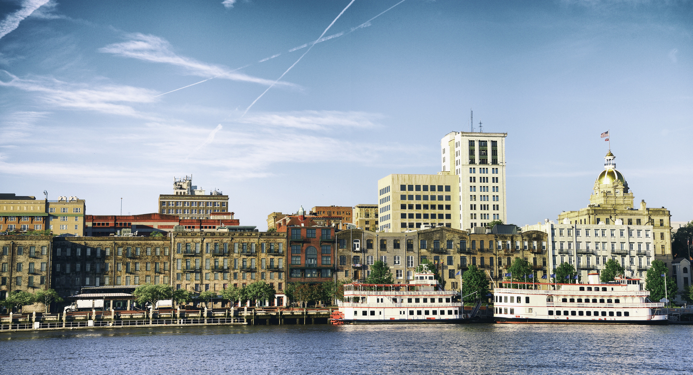 The Savannah riverfront, offering trips that feel luxurious, features historic brick buildings, a tall white tower, a gold-domed city hall, and two riverboats docked on the water.