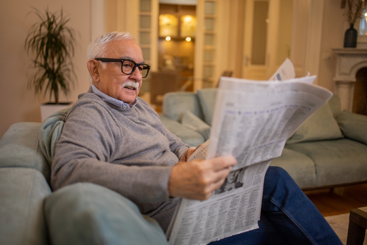 An elderly man with a white mustache and glasses sitting on a sofa while reading a newspaper.
