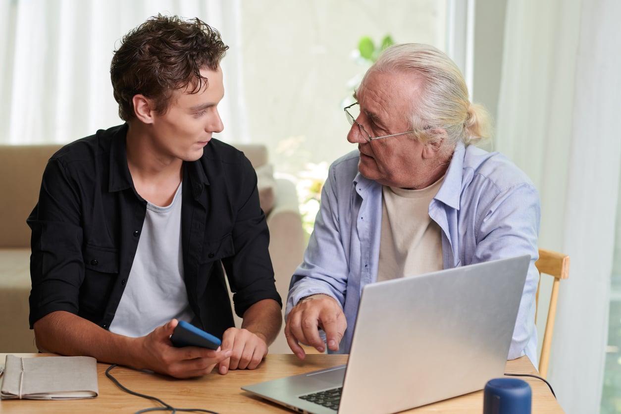 An older man with a ponytail and glasses shows a younger man something on a laptop screen.
