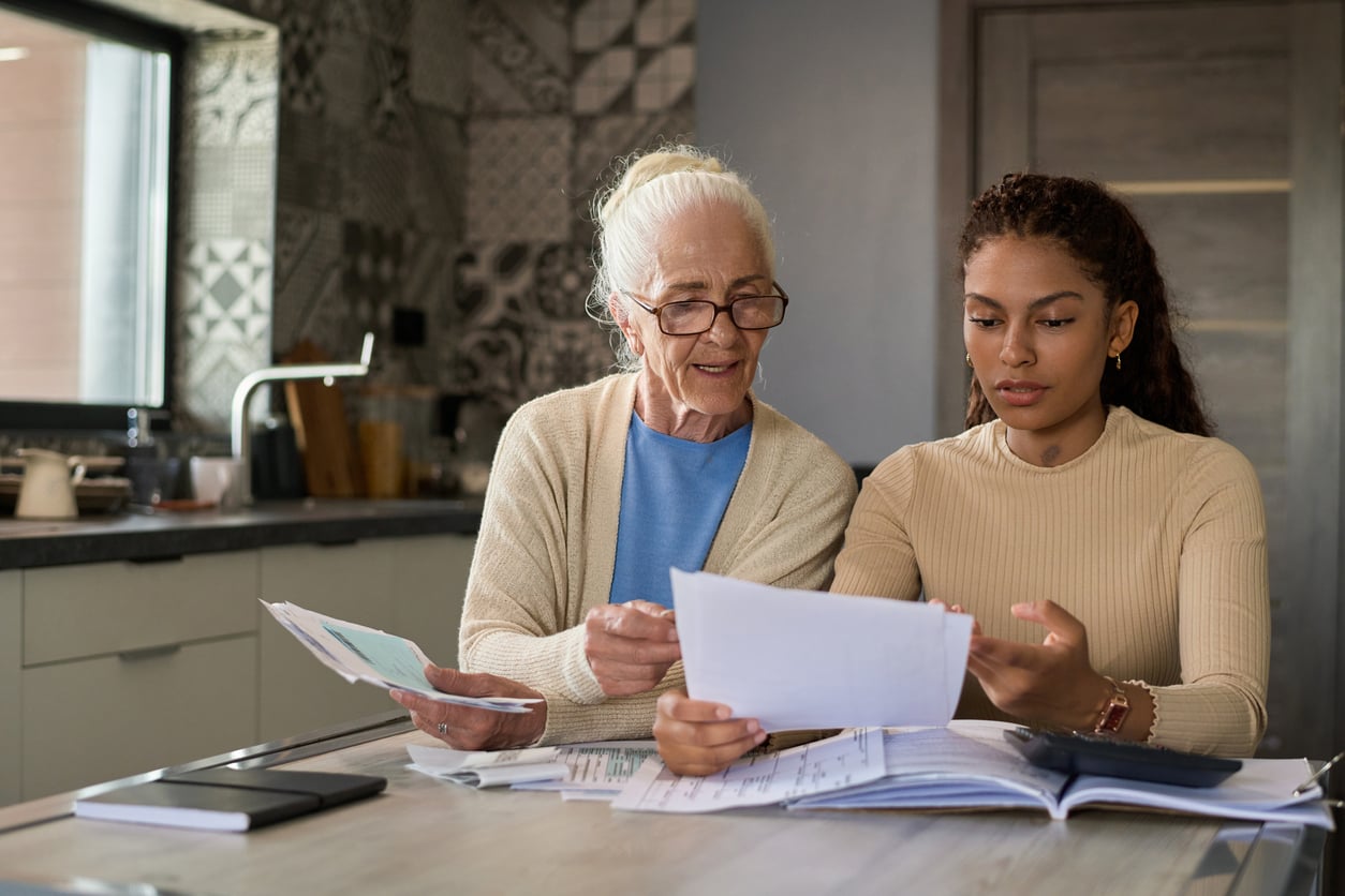 An older woman with glasses and a younger woman sit at a table reviewing paperwork together.