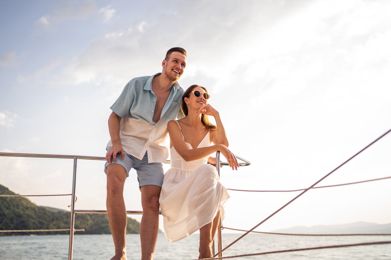 A man and a woman in light summer clothing sit on the railing of a boat, enjoying the cruise entertainment while looking out over the water.