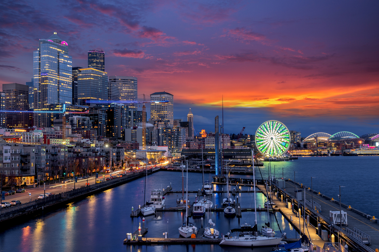 Seattle waterfront at sunset with colorful sky, illuminated skyscrapers, a glowing Ferris wheel, and boats docked at the marina.