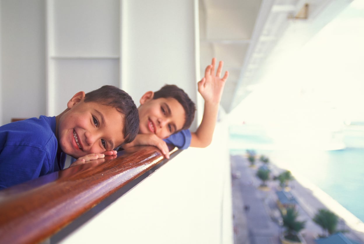 Two boys lean over the cruise ship railing, with one boy waving at the amenities below.