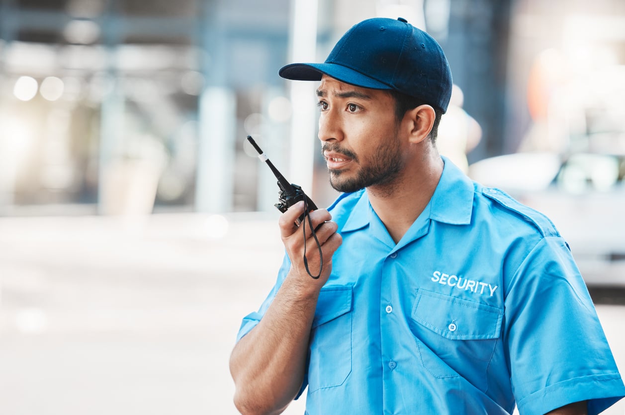 A security guard in a blue uniform and cap speaks into a handheld two-way radio.