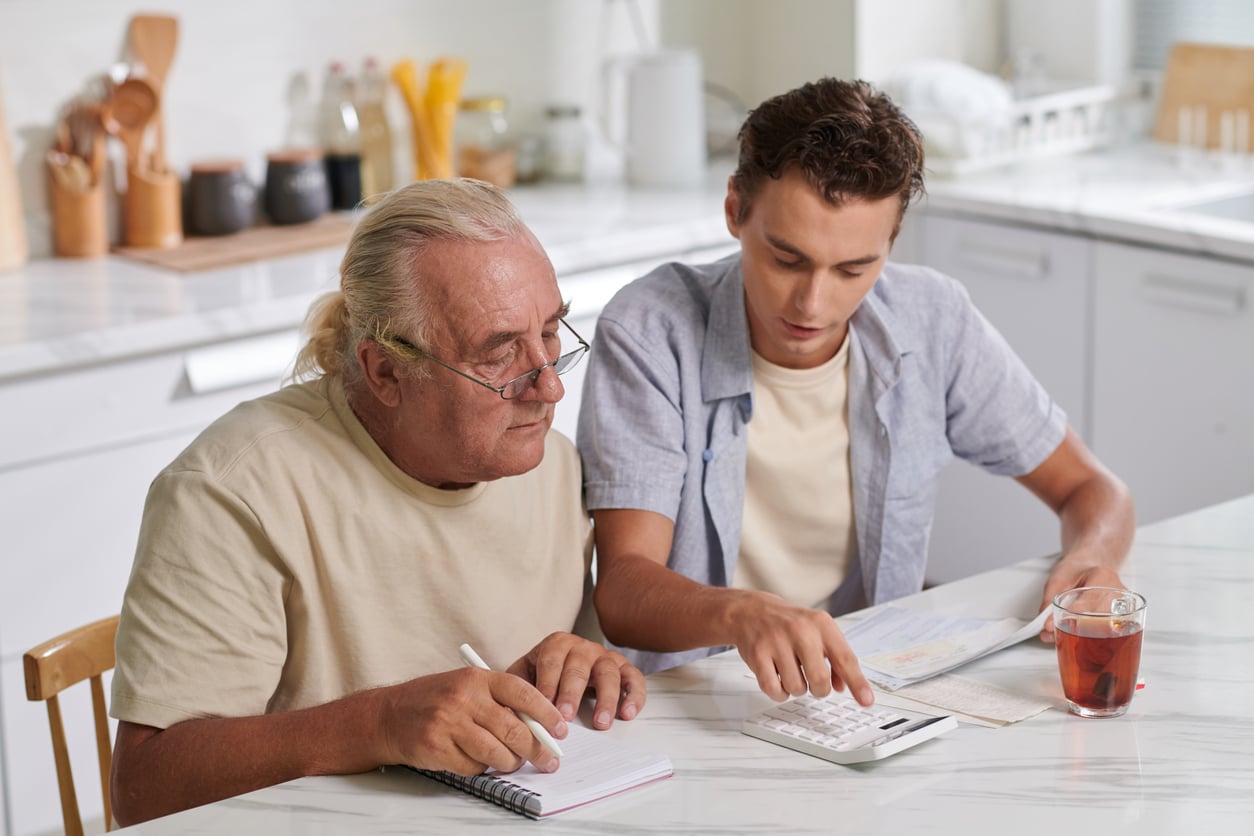 An older man and a young man sit at a kitchen table looking at financial documents and using a calculator.
