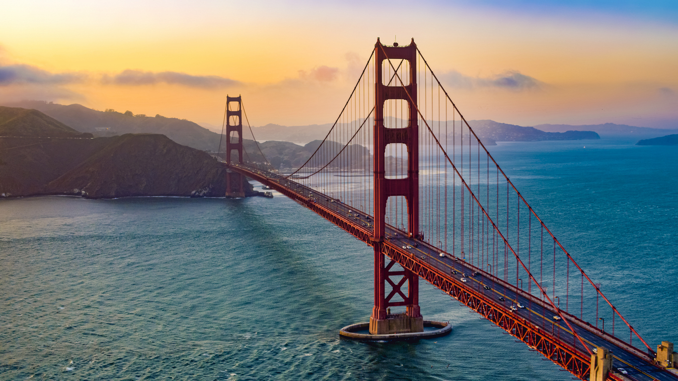 The Golden Gate Bridge stretches over blue water at sunset, with orange and pink skies, distant hills, and a few clouds in the background.