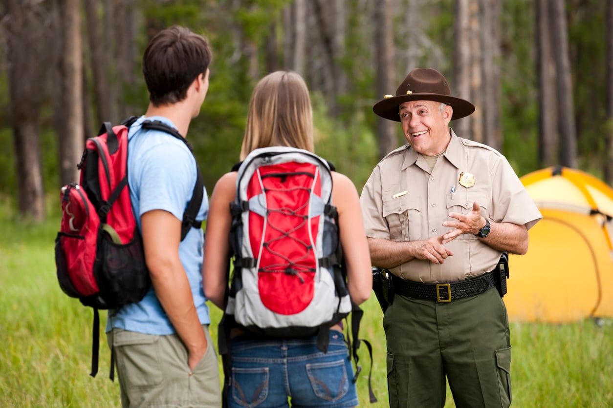 A park ranger speaks to two hikers wearing backpacks in a forest clearing.