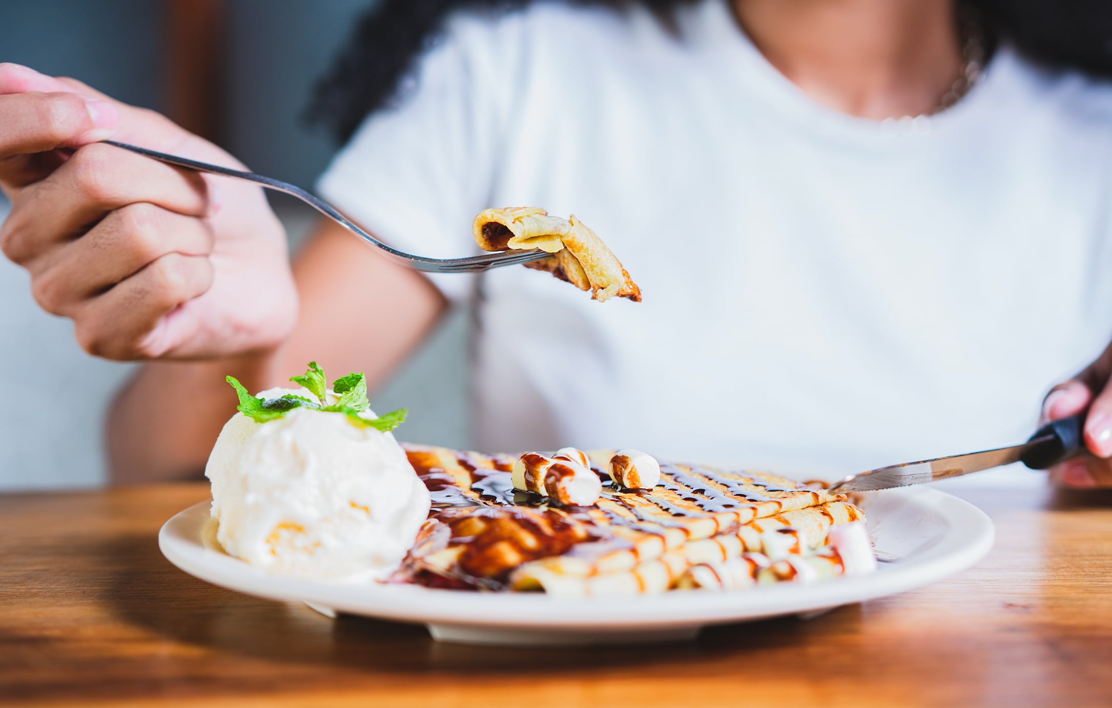 A person uses a fork to lift a piece of a chocolate-drizzled crepe, a delicious breakfast food served with a scoop of ice cream on a white plate.
