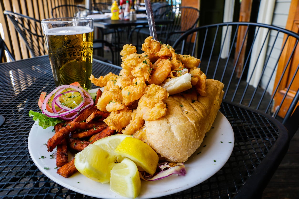 A fried shrimp po’ boy sandwich served with sweet potato fries, lemon wedges, and a glass of beer on a patio table.
