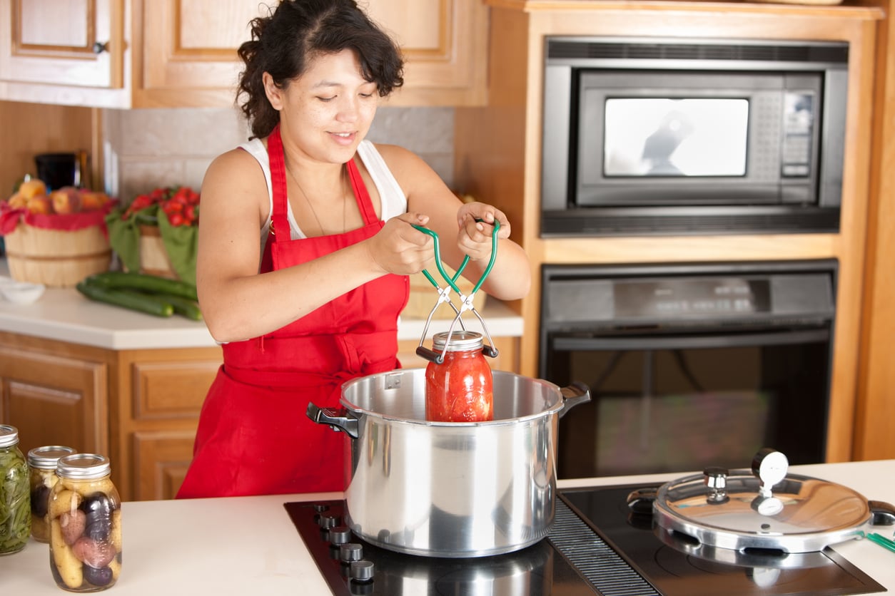 A person wearing a red apron uses canning tongs to lift a jar of tomatoes out of a large pot on a stovetop.