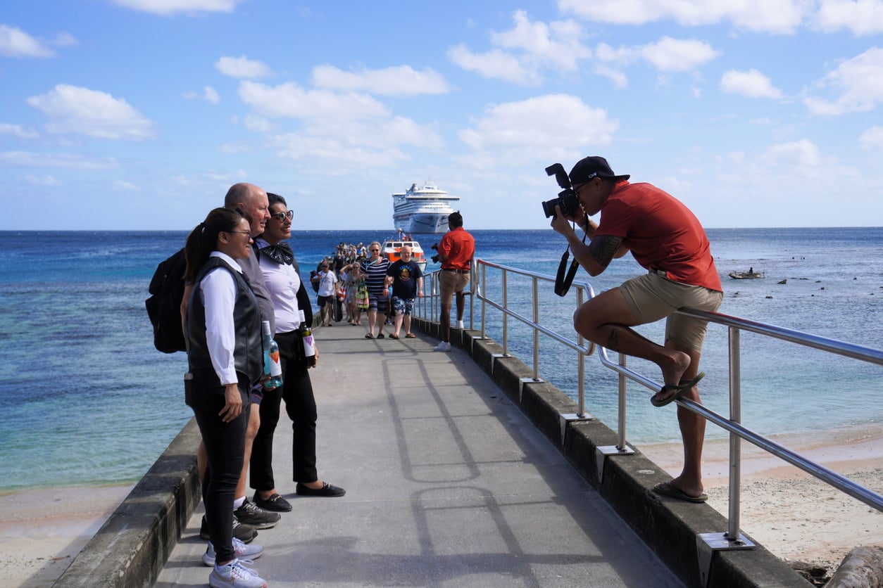A photographer takes a picture of a group of people posing on a concrete pier, with a cruise ship in the background.