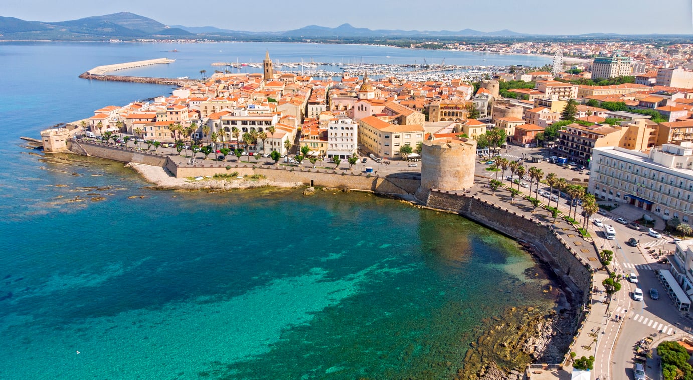 Aerial view of the fortified historic center of Alghero, Sardinia, featuring stone walls, a coastal tower, and a harbor filled with boats along the turquoise Mediterranean Sea.