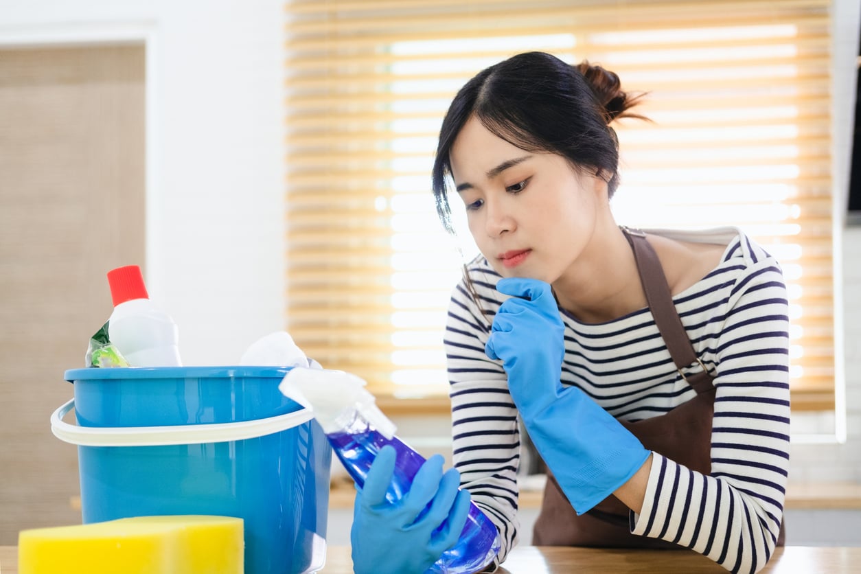 A woman wearing blue rubber gloves and an apron looks thoughtfully at a bottle of blue cleaning spray next to a bucket of cleaning supplies.