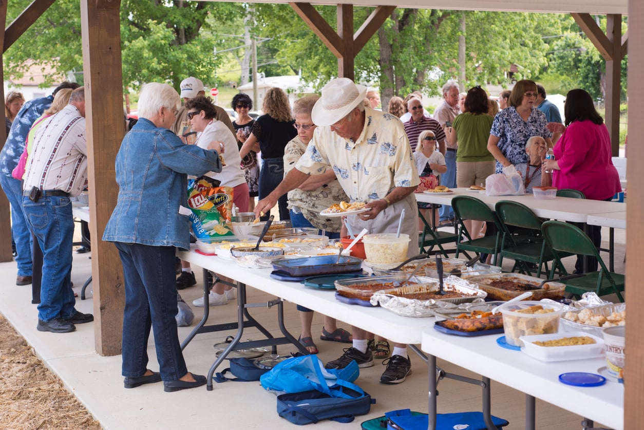 People serving themselves food from a buffet table at an outdoor park pavilion.