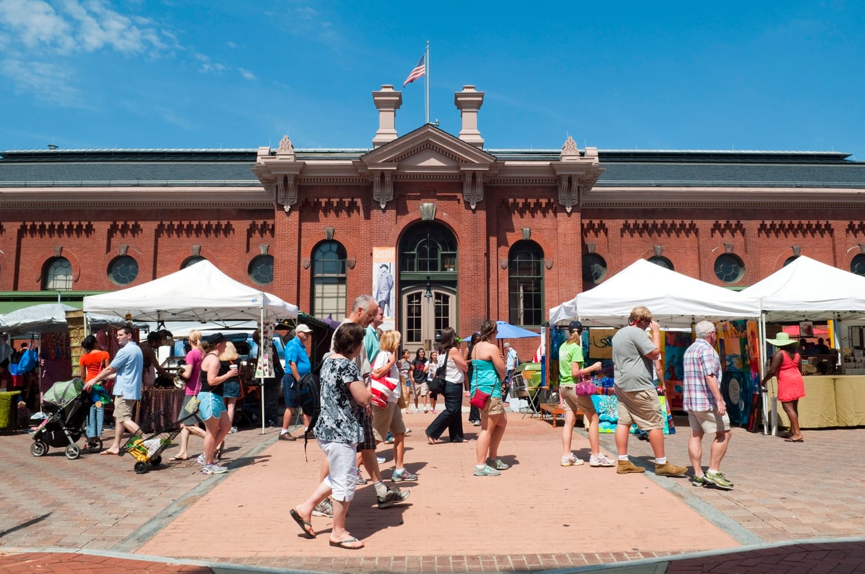 People browse white vendor tents in a plaza in front of a historic brick market building under a clear blue sky.