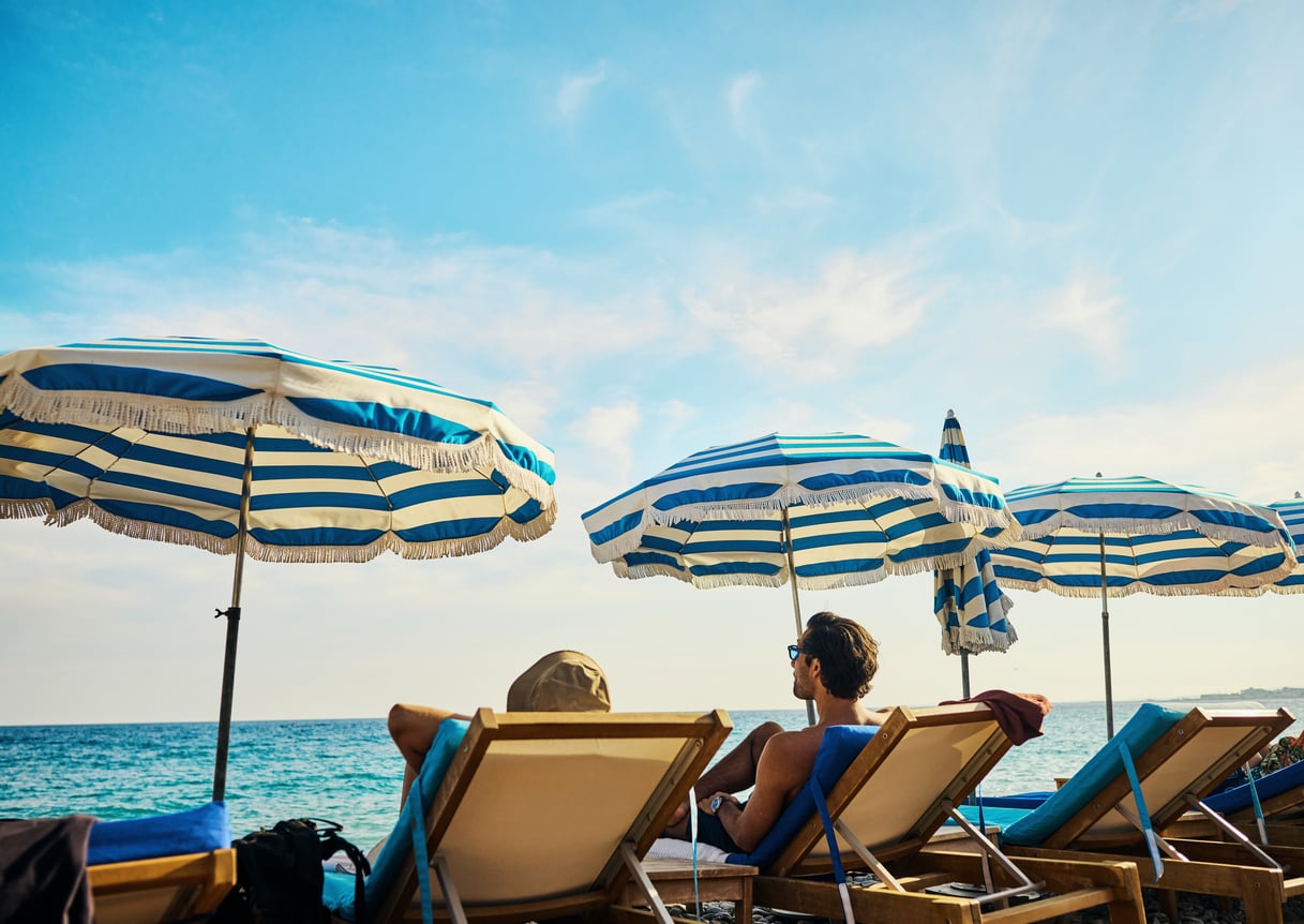Two people lounging in beach chairs under blue and white striped umbrellas overlooking the sea.