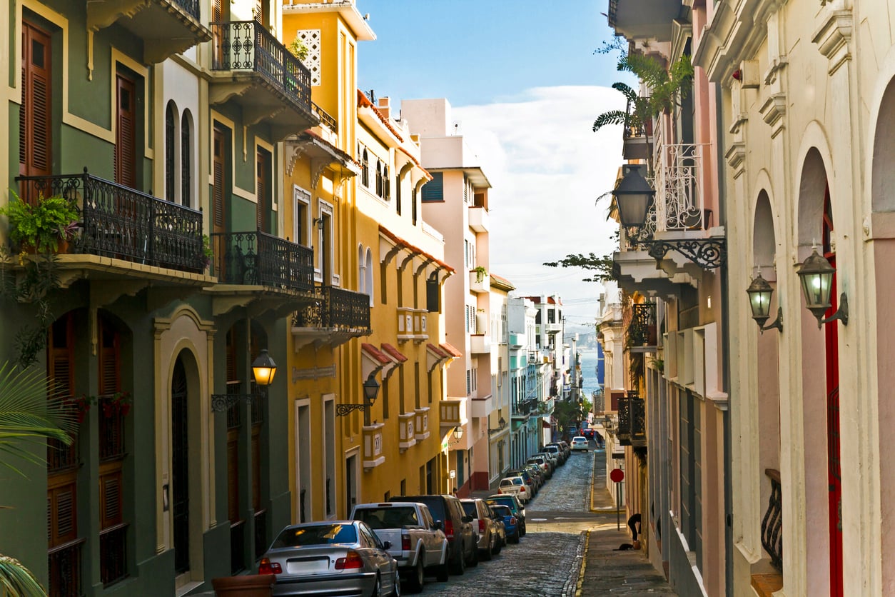 A narrow cobblestone street lined with colorful colonial-style buildings, wrought-iron balconies, lanterns, and parked cars, leads down toward the sea under a bright blue sky.