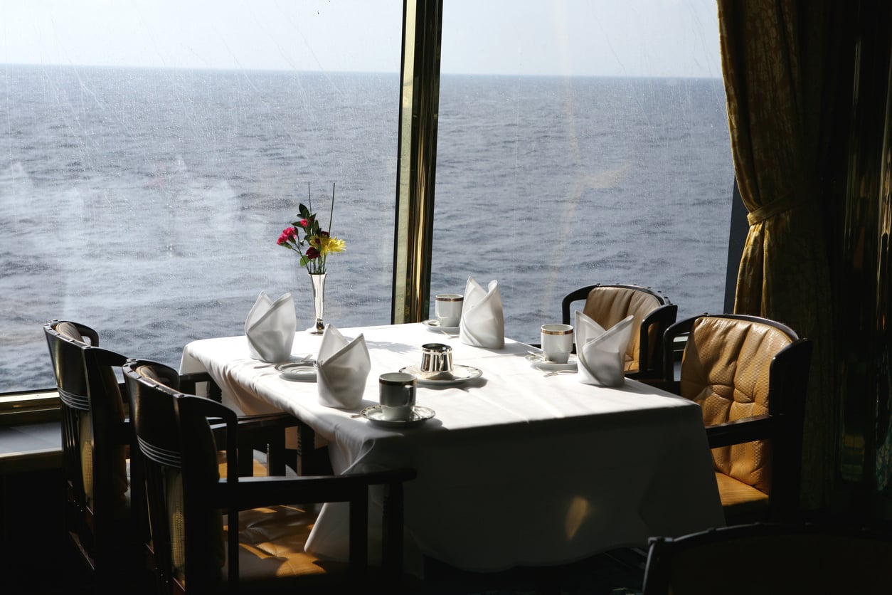 A dining table set for four with white napkins and cups next to a large window overlooking the ocean.