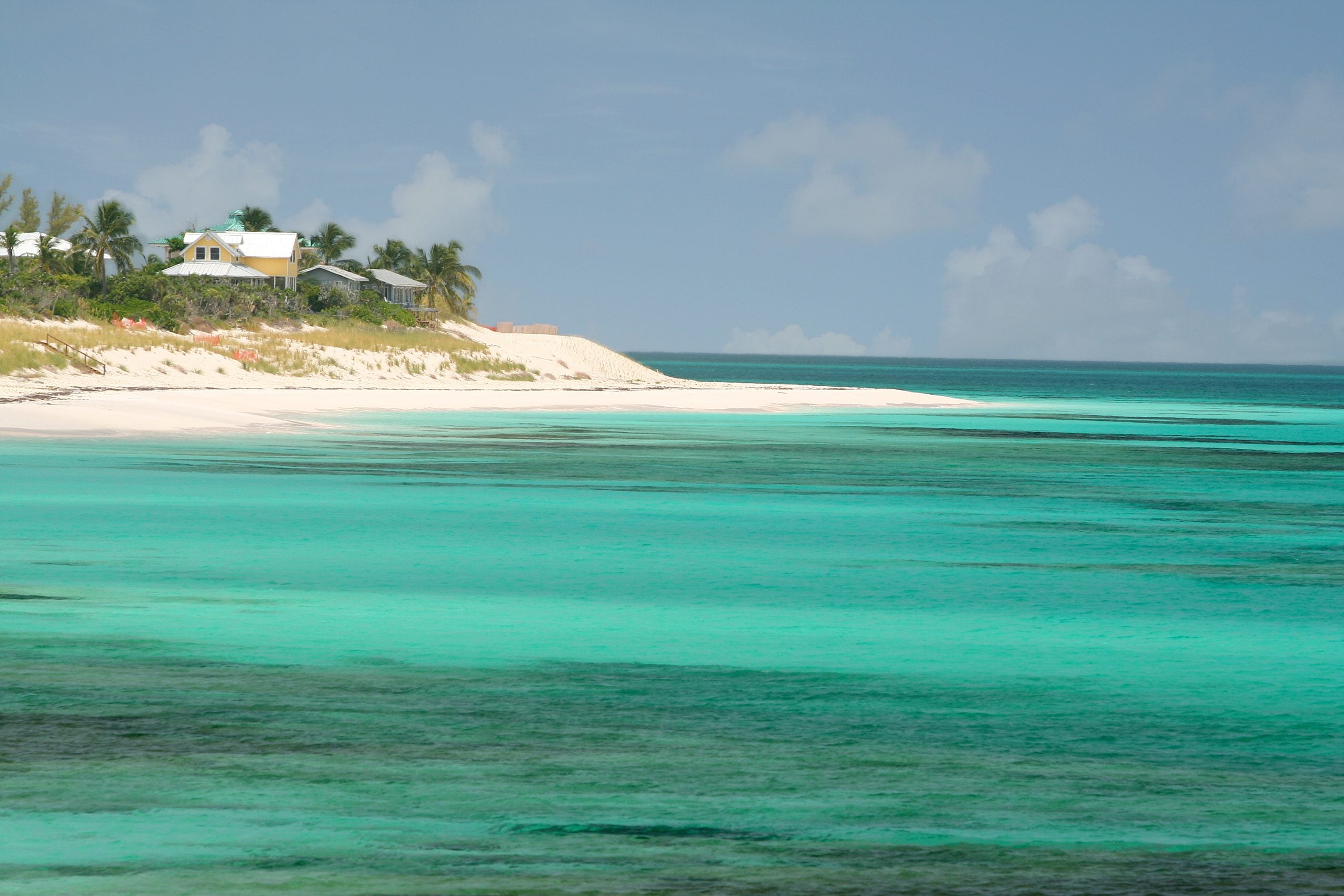 A yellow house sits on a sandy shore overlooking turquoise water.