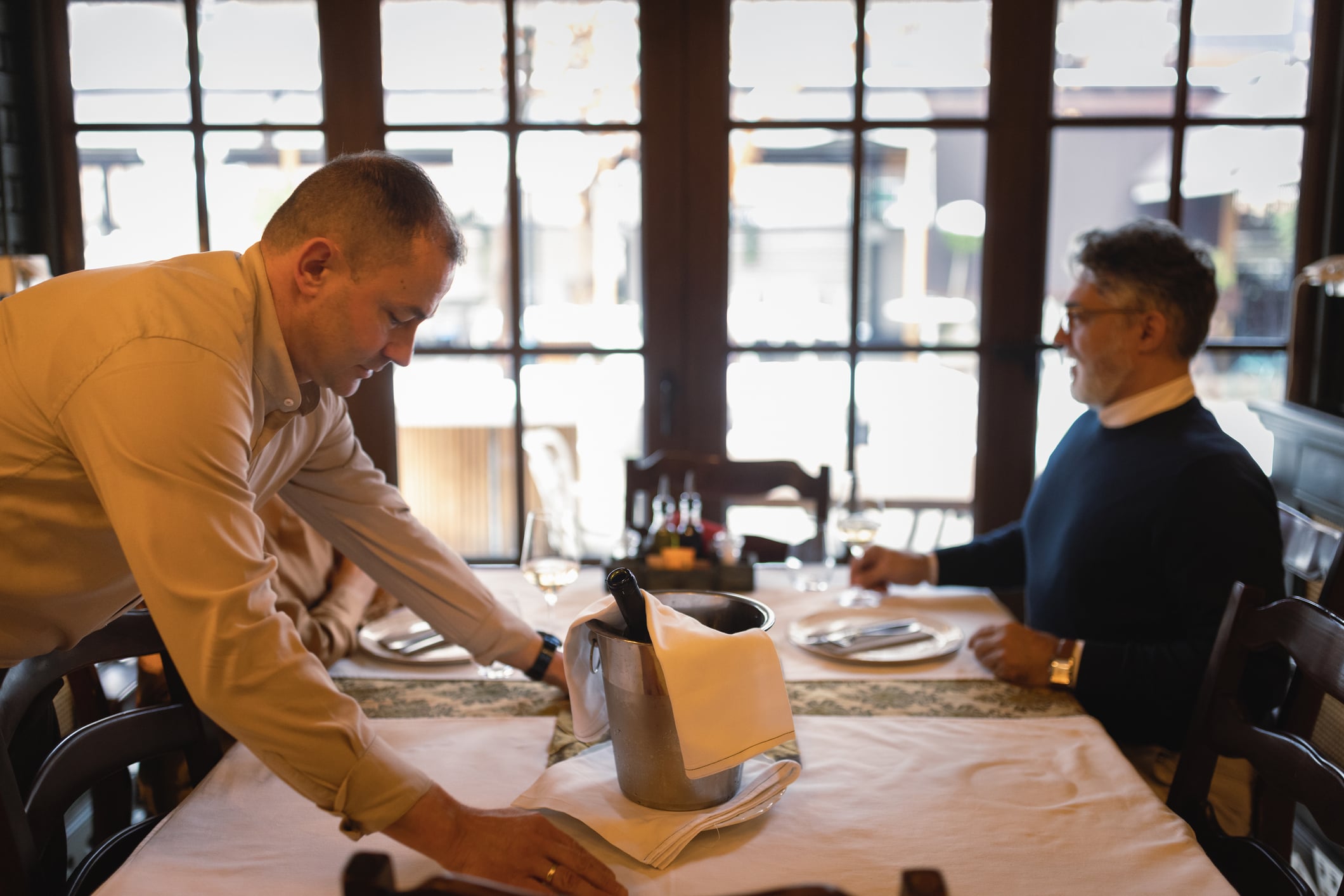 A waiter in a tan shirt sets a table for a customer sitting across from him, with a champagne bucket placed in the center of the table.