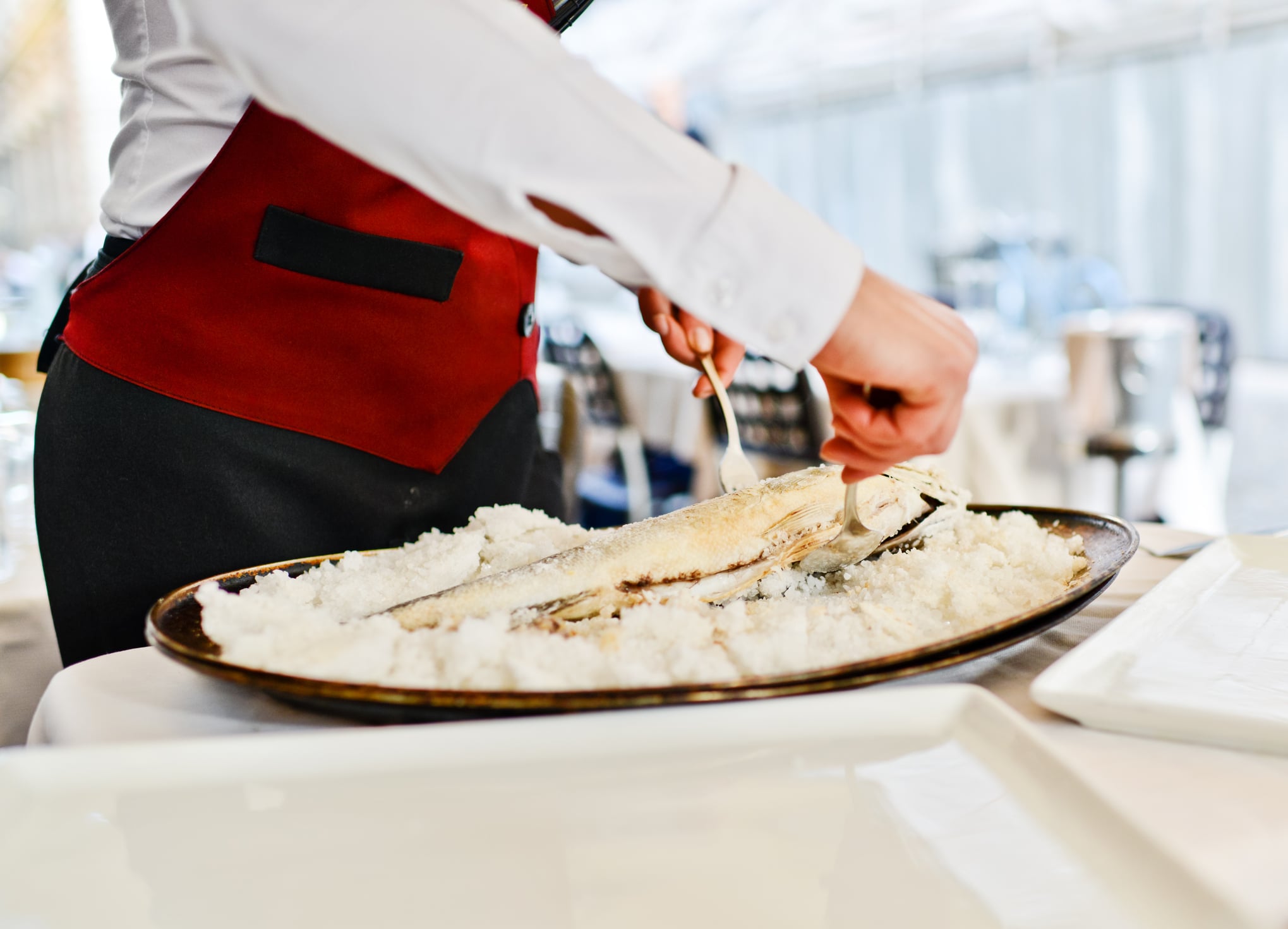 A server in a white shirt and red apron uses a fork and spoon to de-bone a whole fish baked in a salt crust on a platter.