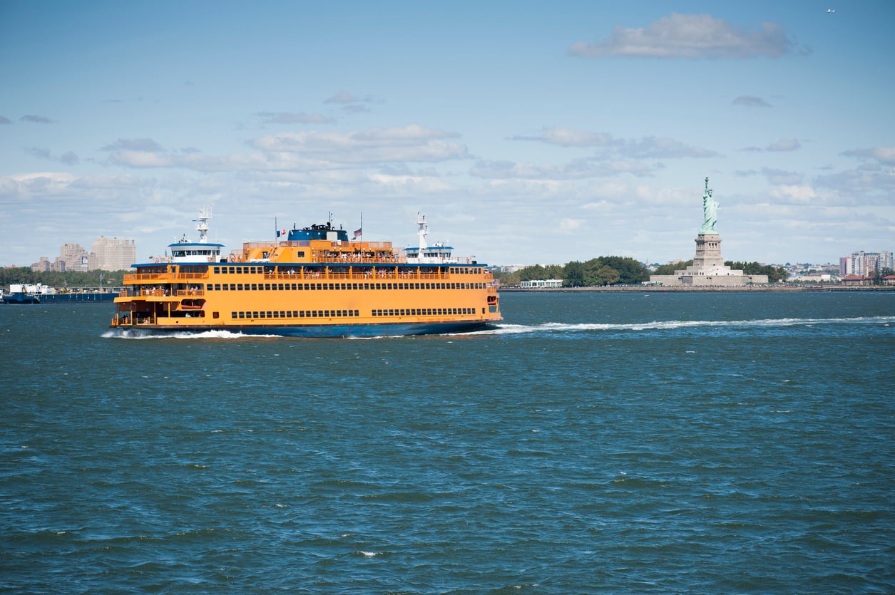 things tourists pay extra for A bright orange Staten Island Ferry boat crossing the water with the Statue of Liberty in the distance.