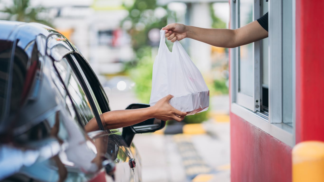 A drive-thru worker hands a white plastic takeout bag to a driver through a service window at one of the fast-food restaurants.