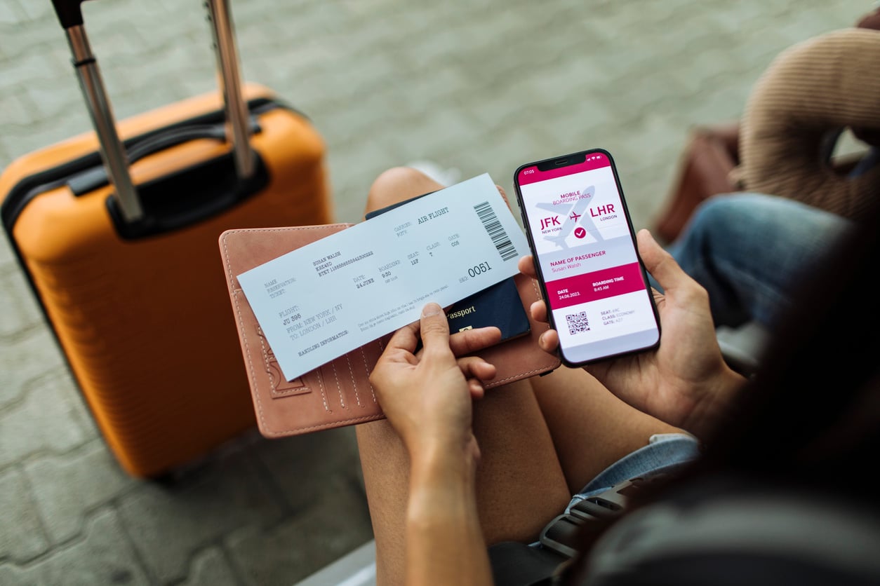 vacation mistakes A traveler holds a paper airline ticket and a smartphone displaying a digital boarding pass for a flight from New York to London, with an orange suitcase nearby.