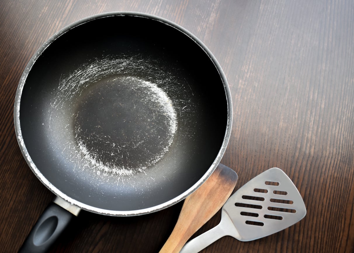 A worn non-stick frying pan with a scratched surface, accompanied by a wooden spatula and a metal slotted turner on a wooden table.