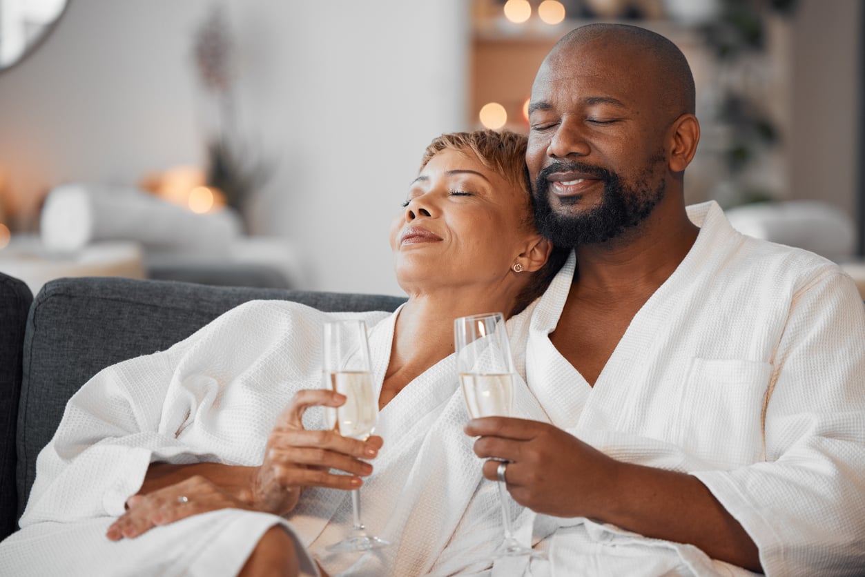 A Black couple in white bathrobes sitting on a sofa with their eyes closed, each holding a glass of champagne.