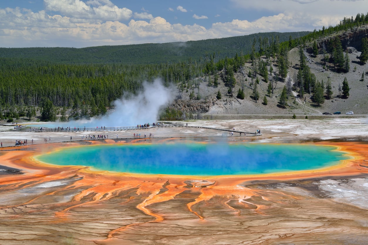 The Grand Prismatic Spring with its vibrant blue center and orange-rimmed terraces, surrounded by a forest and a wooden boardwalk with people walking on it.