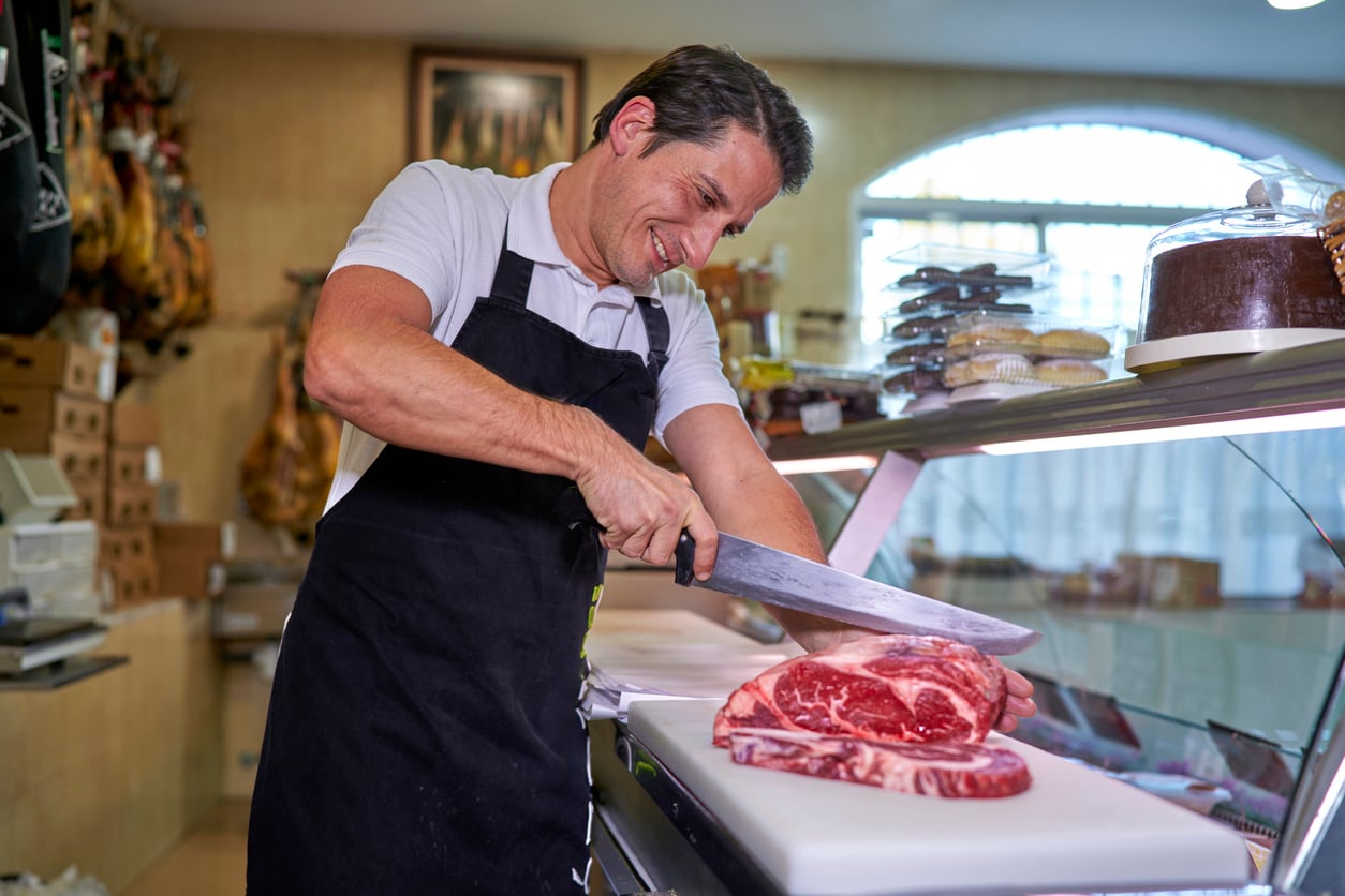 A smiling butcher in a black apron slices a raw ribeye steak on a cutting board behind a deli counter.
