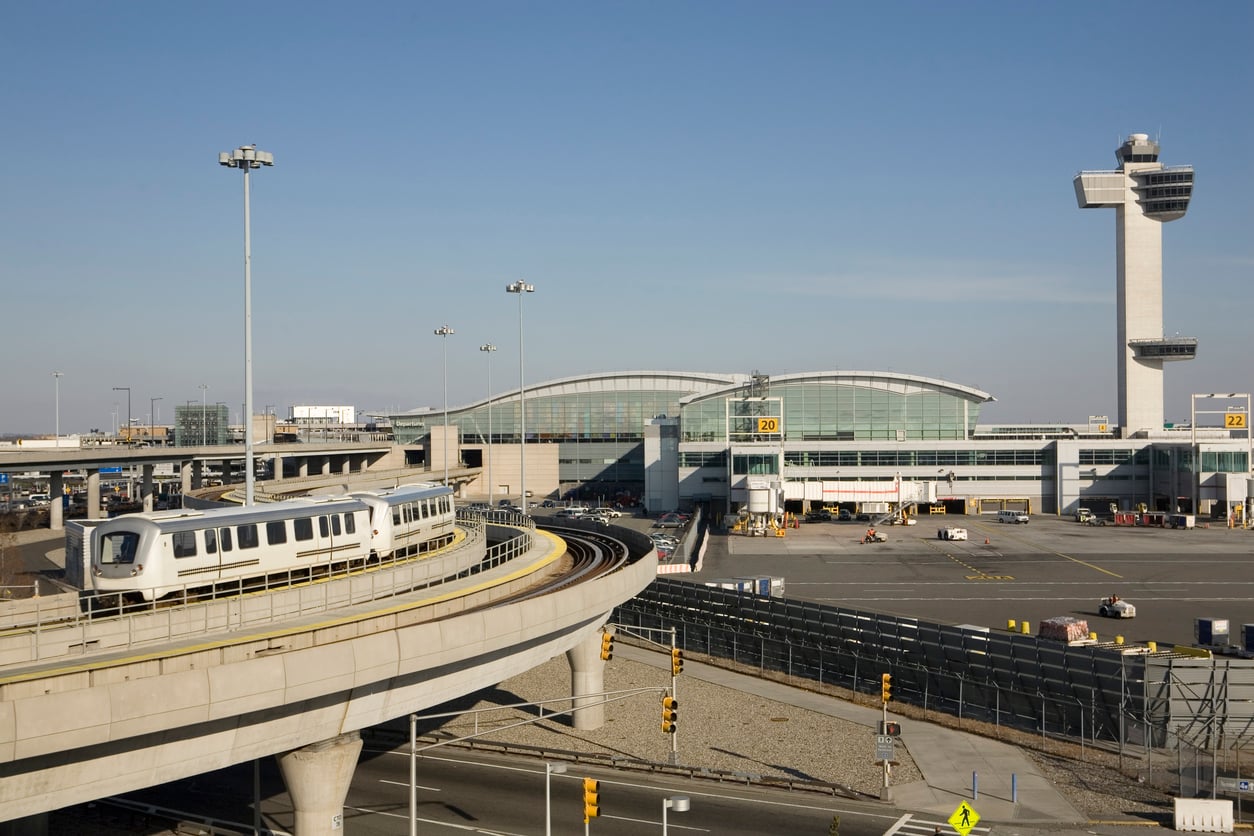 An airport train curves on an elevated track near a terminal building and air traffic control tower.