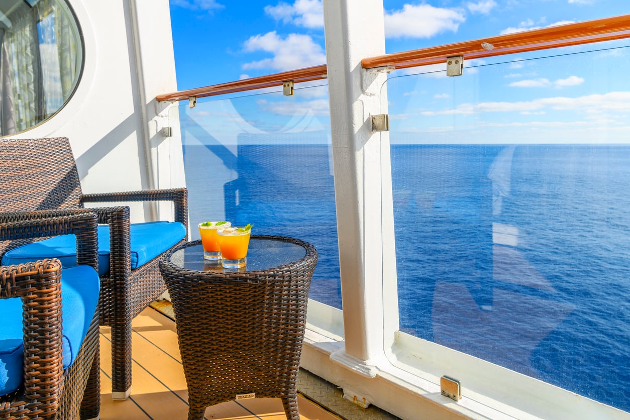 Two orange cocktails on a round wicker table between two wicker chairs on a cruise ship balcony overlooking the ocean.