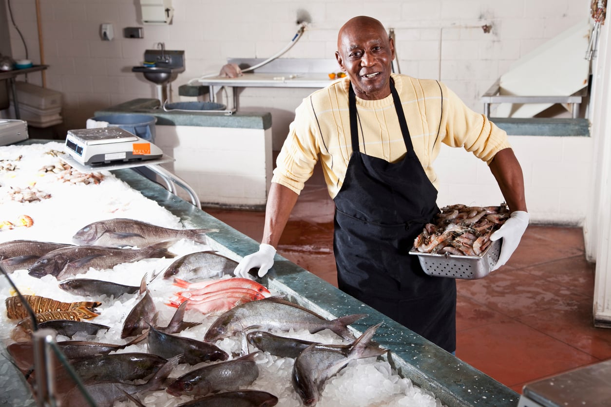 A smiling fishmonger wearing a yellow sweater and black apron holds a metal container of raw shrimp over a bed of ice filled with fresh fish.