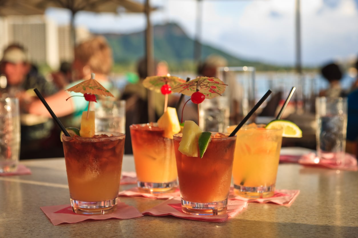 Four tropical cocktails with pineapple, lime, and paper umbrellas sit on a table overlooking a waterfront view, where cruise ship charges may apply.