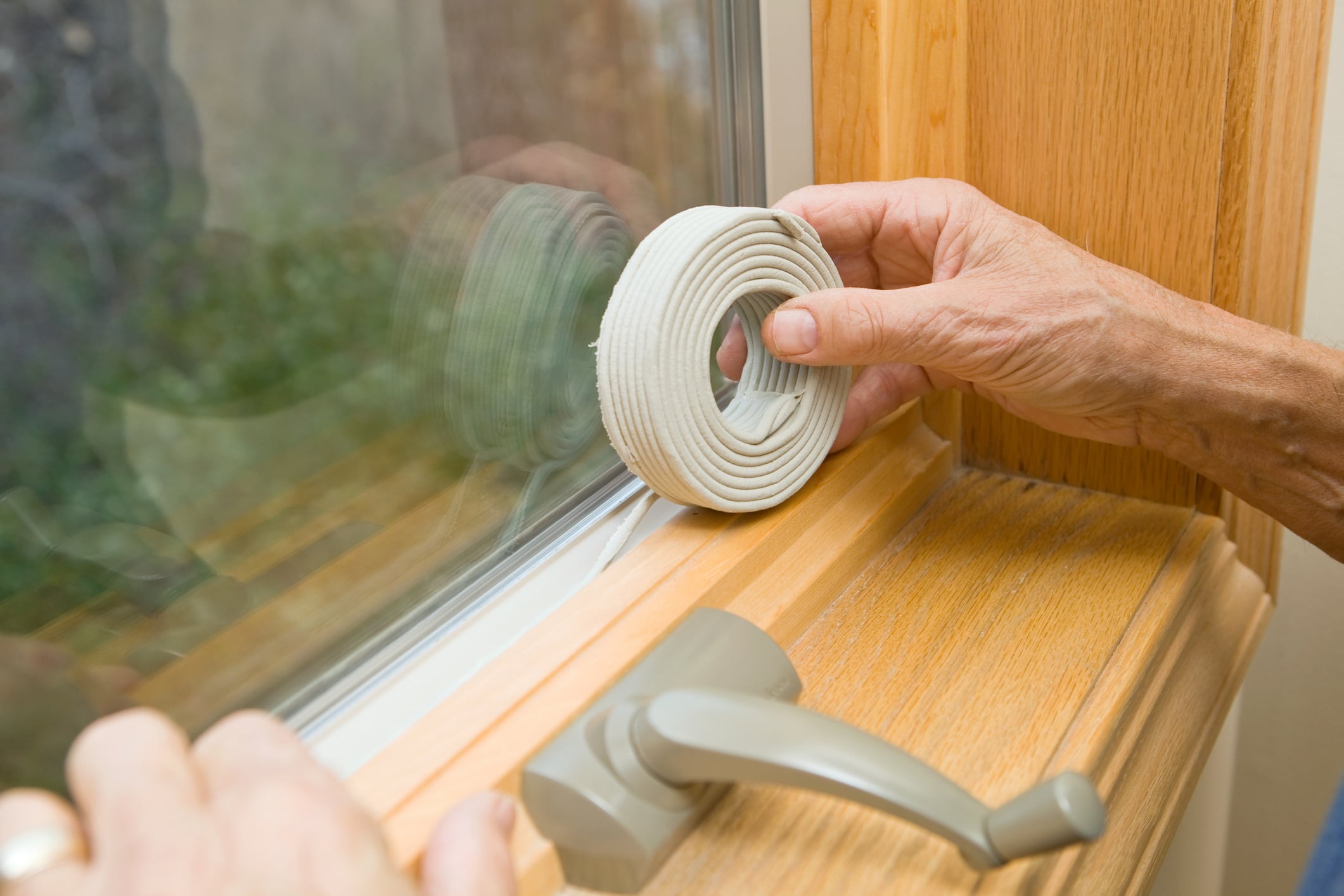 A person applying a roll of adhesive foam weatherstripping to the frame of a wooden window.