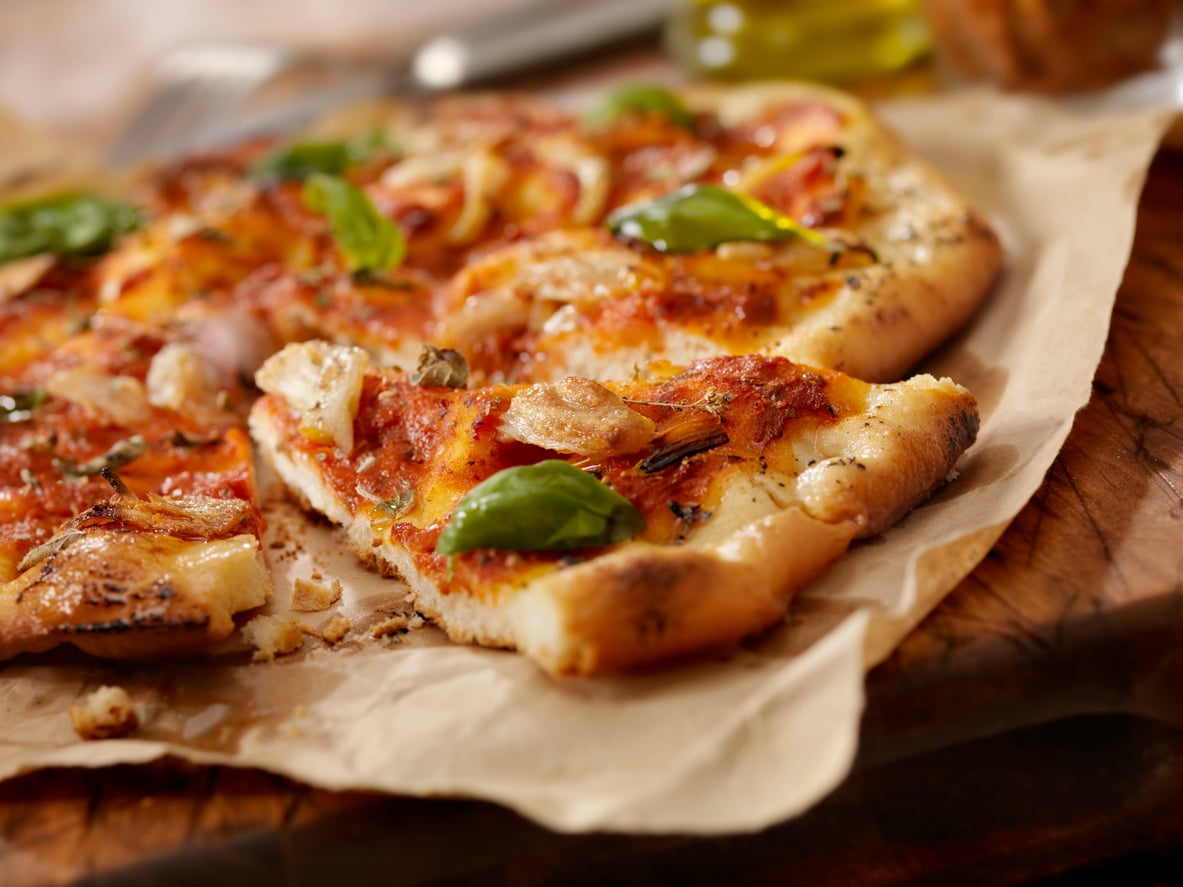 A slice of tomato pizza topped with roasted garlic cloves and fresh basil leaves on parchment paper.