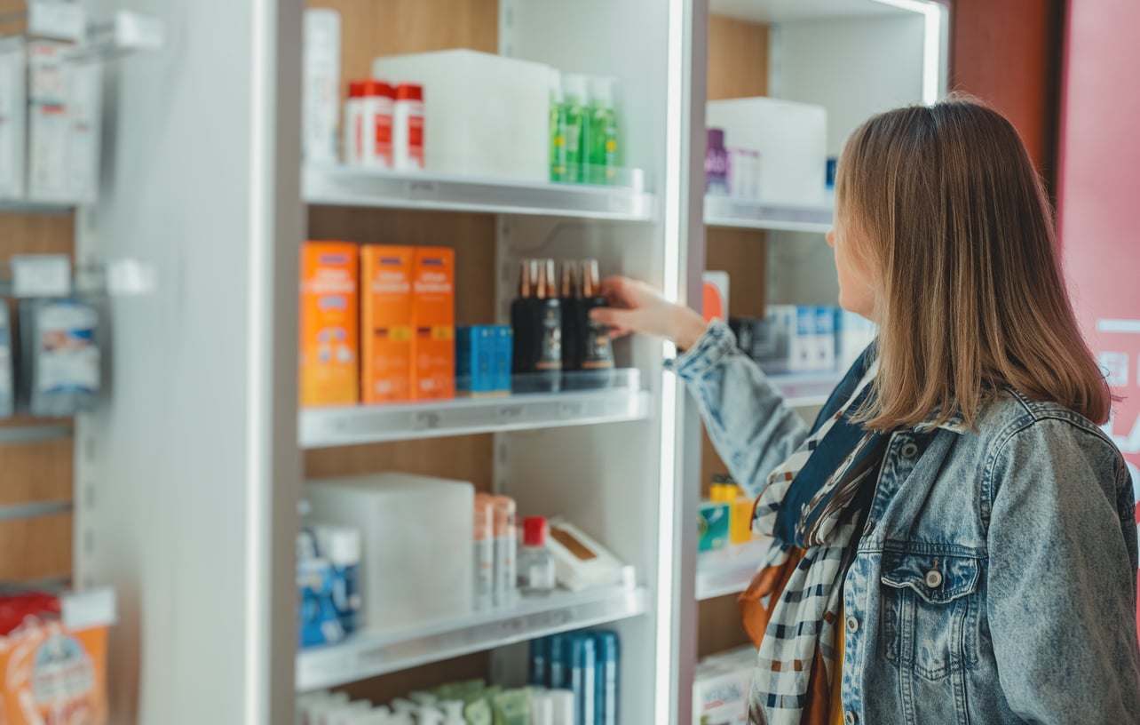 A woman in a denim jacket reaches for a product on a store shelf filled with various bottles and packages.